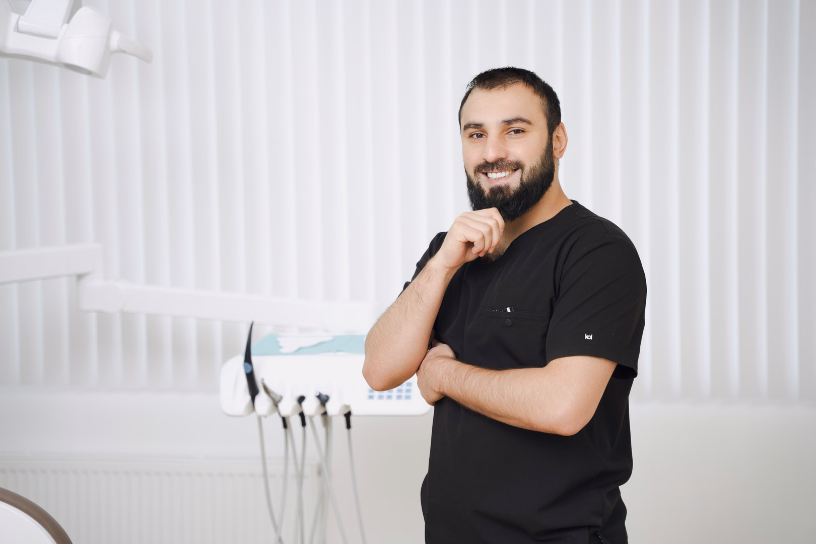 Бородатый сотрудник Simpla Dental улыбается и держит руку у подбородка рядом со стоматологической установкой