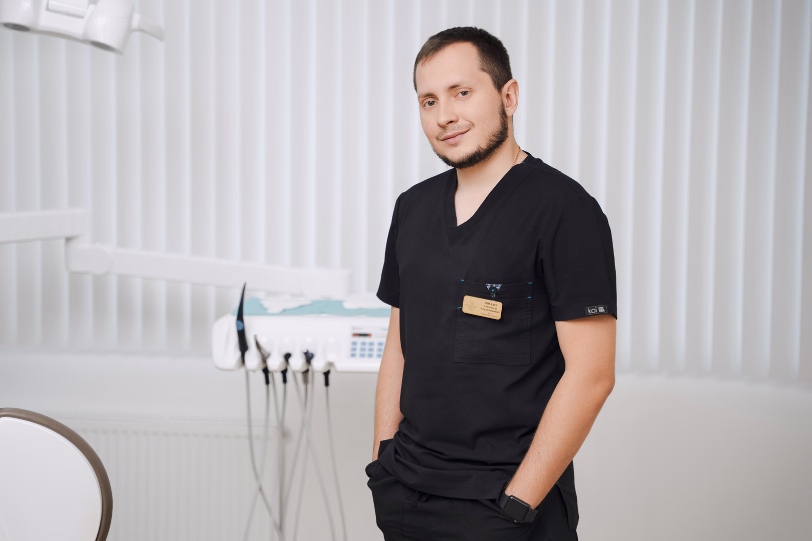 Сотрудник Simpla Dental в чёрной форме стоит вполоборота с рукой в кармане на фоне жалюзи