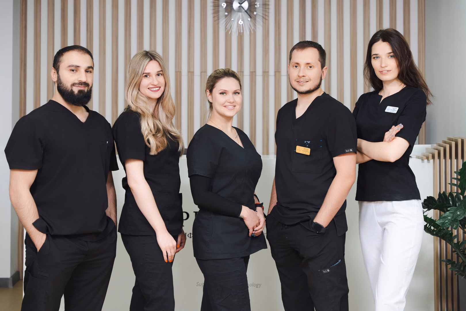 Пять сотрудников Simpla Dental позируют вместе на фоне деревянных реек и светлых жалюзи