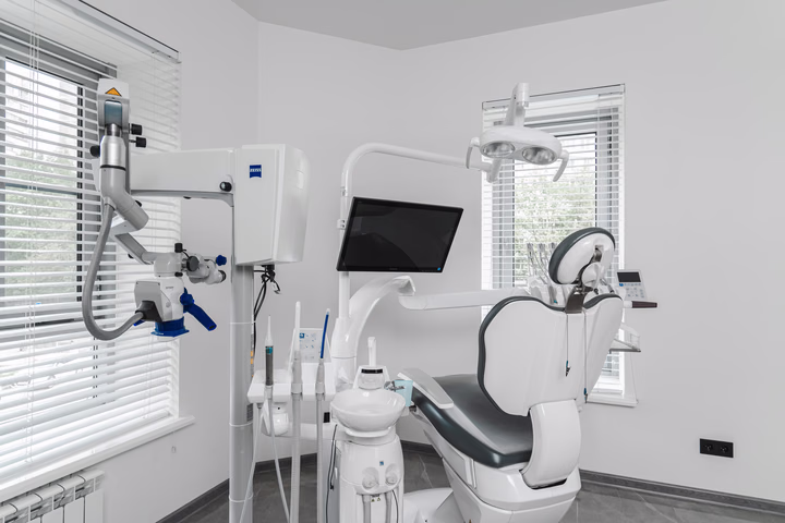 Тёмное стоматологическое кресло и микроскоп с синими ручками у окна в Dental Smile