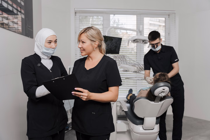 Две сотрудницы Dental Smile с планшетом для бумаг на фоне приёма пациента в кресле