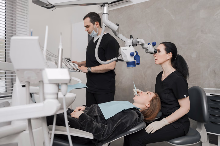 Сотрудница Dental Smile работает у микроскопа, рядом коллега у блока инструментов и пациентка