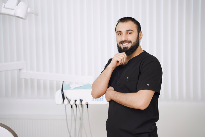 Бородатый сотрудник Simpla Dental улыбается и держит руку у подбородка рядом со стоматологической установкой