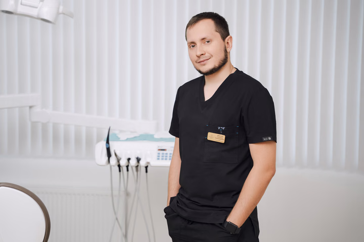 Сотрудник Simpla Dental в чёрной форме стоит вполоборота с рукой в кармане на фоне жалюзи