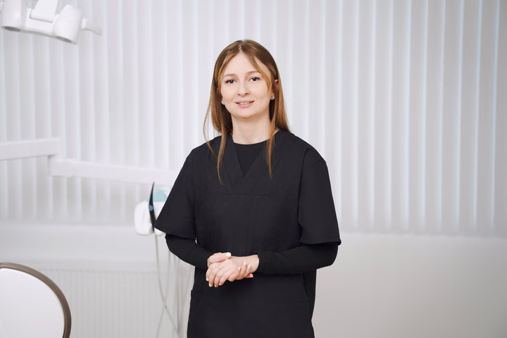 Сотрудница Simpla Dental с прямыми рыжими волосами держит руки перед собой на фоне светлых жалюзи