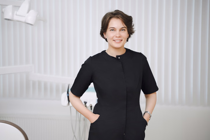 Улыбающаяся сотрудница Simpla Dental с короткой стрижкой стоит, убрав руки в карманы чёрной формы