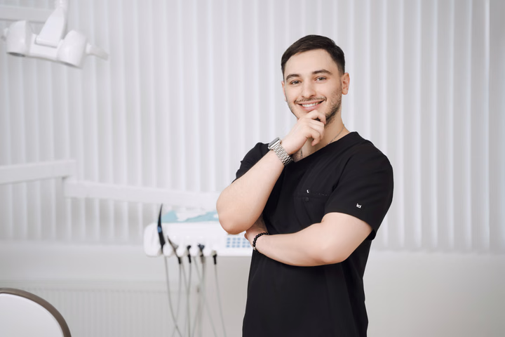 Улыбающийся сотрудник Simpla Dental держит одну руку у подбородка, другую сложил на груди