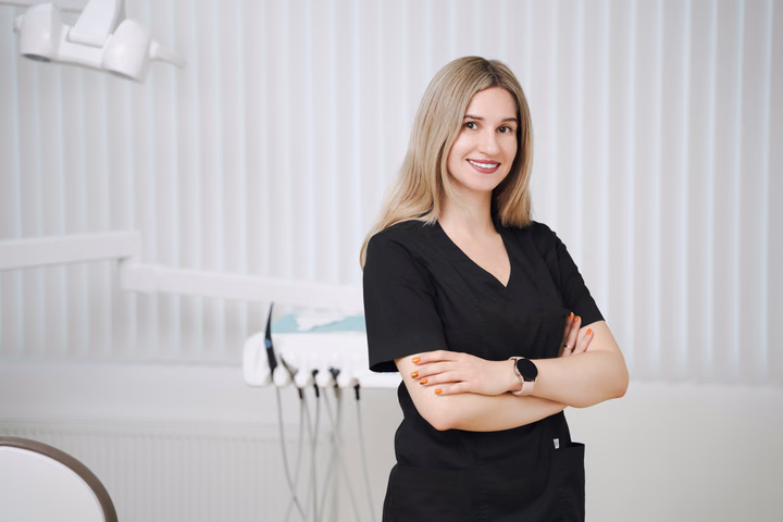 Сотрудница Simpla Dental со светлыми волосами улыбается, скрестив руки на фоне белых жалюзи