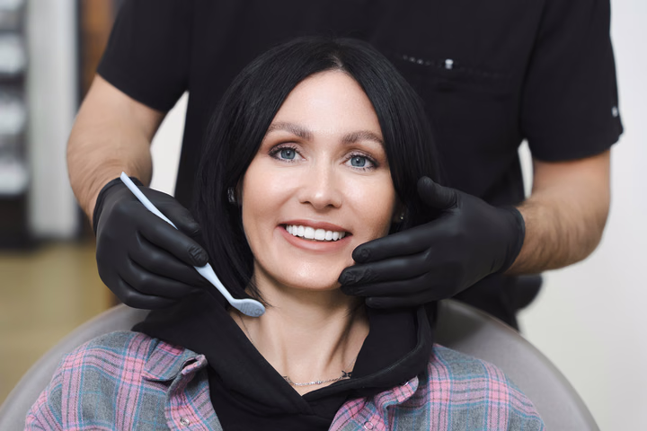 Пациентка Simpla Dental с тёмными волосами в кресле, рядом с лицом зеркало и руки в чёрных перчатках