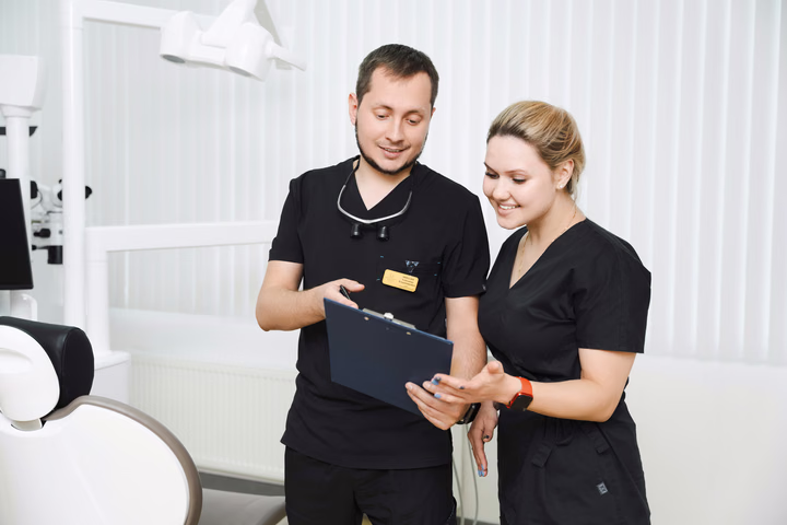 Сотрудник и сотрудница Simpla Dental улыбаются, рассматривая записи на синем планшете для бумаг