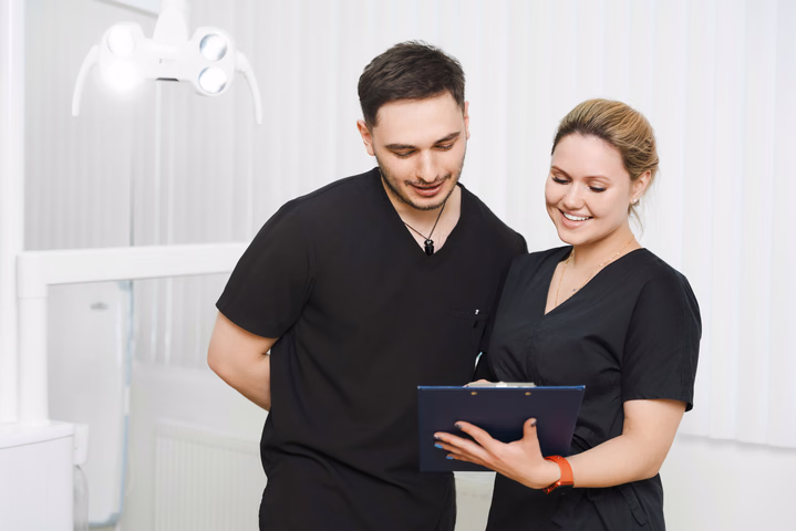 Сотрудник Simpla Dental и улыбающаяся коллега рассматривают синий планшет с бумагами, вид по пояс