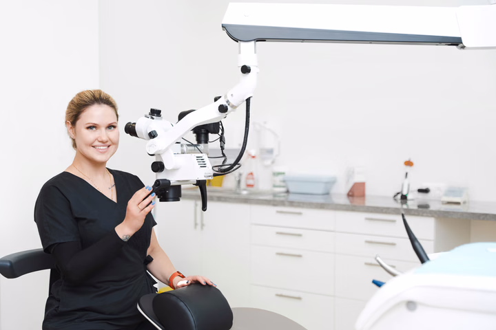 Улыбающаяся сотрудница Simpla Dental сидит рядом с микроскопом и держится за его корпус