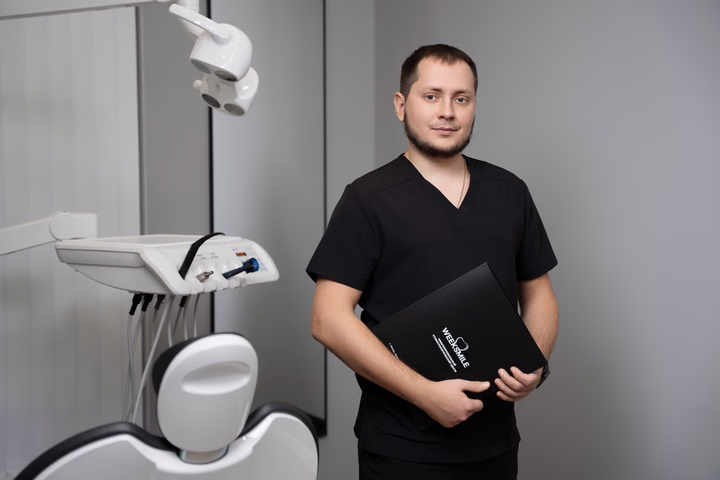 Мужчина в чёрной форме с закрытой папкой у стоматологического кресла Weeksmile