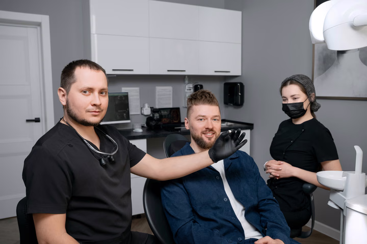 Стоматолог Weeksmile жестом обращается к пациенту в кресле, рядом стоит ассистент